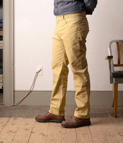 Indoors, a person wears a blue shirt, brown shoes, and Best Made Co. Standard 5-Pocket Pant M2 Khaki on a wooden floor near a wall outlet, with part of a chair visible on the right.