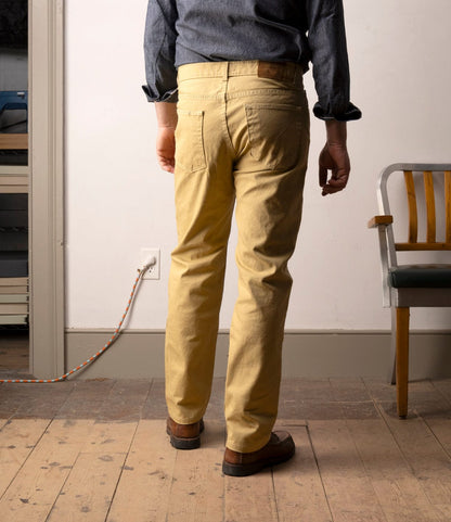 A person stands indoors on a wooden floor, facing away, wearing Best Made Co. Standard 5-Pocket Pant M2 Khaki, a blue shirt, and brown shoes. An empty chair and a plugged-in extension cord are visible in the background.