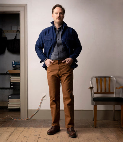 A man stands indoors on a wooden floor, wearing Best Made Co.'s Standard 5-Pocket Pant M2 Field Brown with a blue jacket and gray shirt. Behind him are a wooden chair and shelves with books and bags against a light wall.
