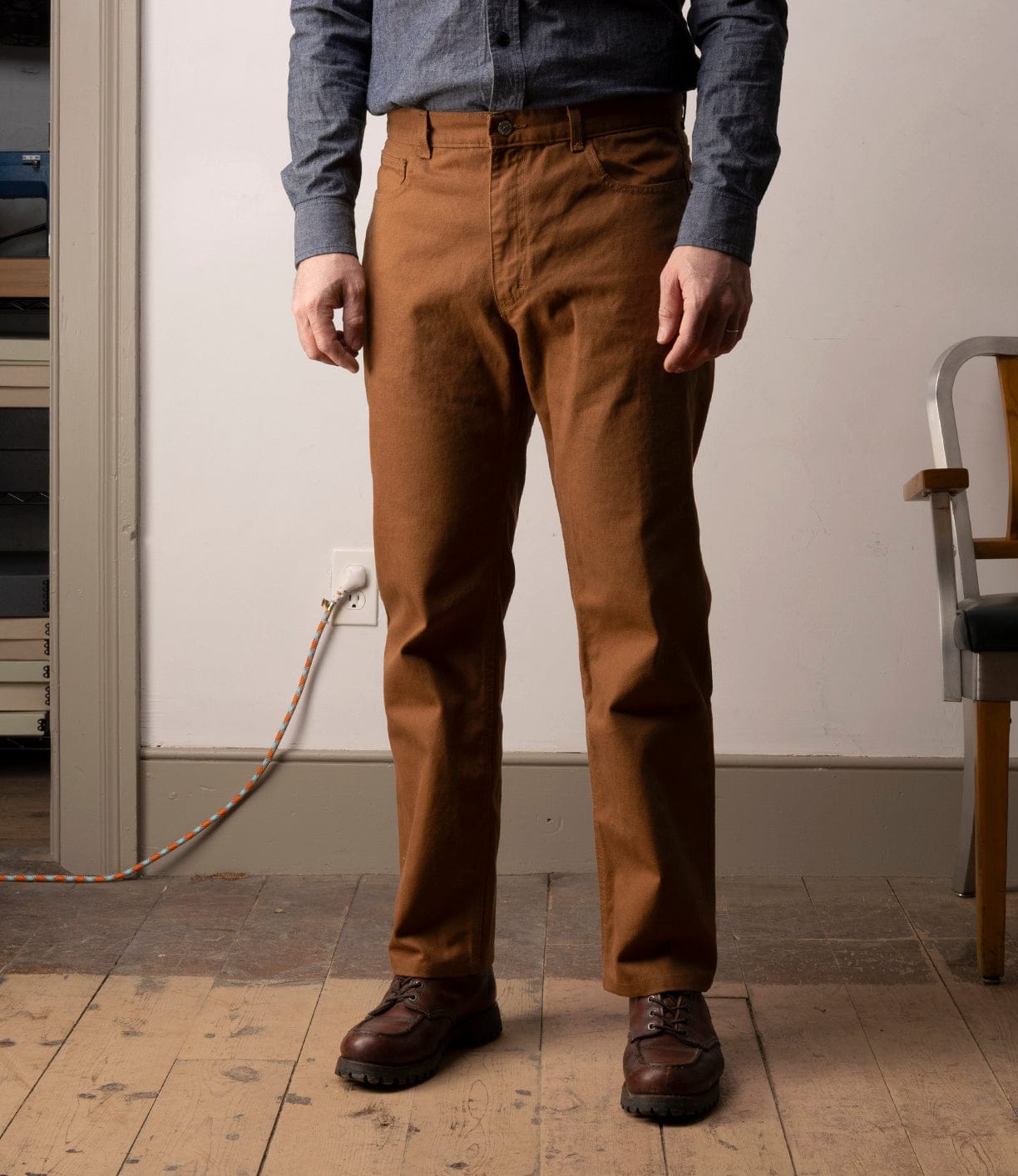 A person wearing Best Made Co Standard 5-Pocket Pant M2 Field Brown, a blue long-sleeve shirt, and brown shoes stands indoors on a wooden floor near a chair and wall with power outlet; their face is not visible.