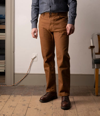 A person stands indoors on a wooden floor, showing only their lower half. They wear Best Made Co.'s Standard 5-Pocket Pant M2 Field Brown with brown shoes, near a gray wall socket and a chair.