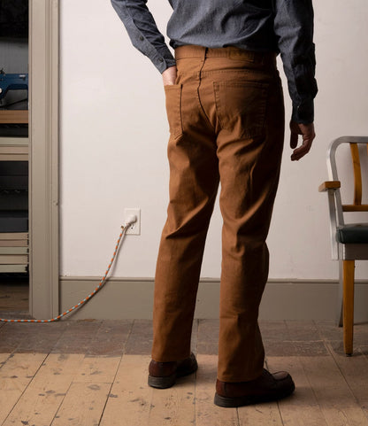 A person stands indoors with their back to the camera, wearing Best Made Co.'s Standard 5-Pocket Pant M2 Field Brown, a blue shirt, and brown shoes. The room has a wooden floor, a chair, and an orange extension cord plugged into the wall.