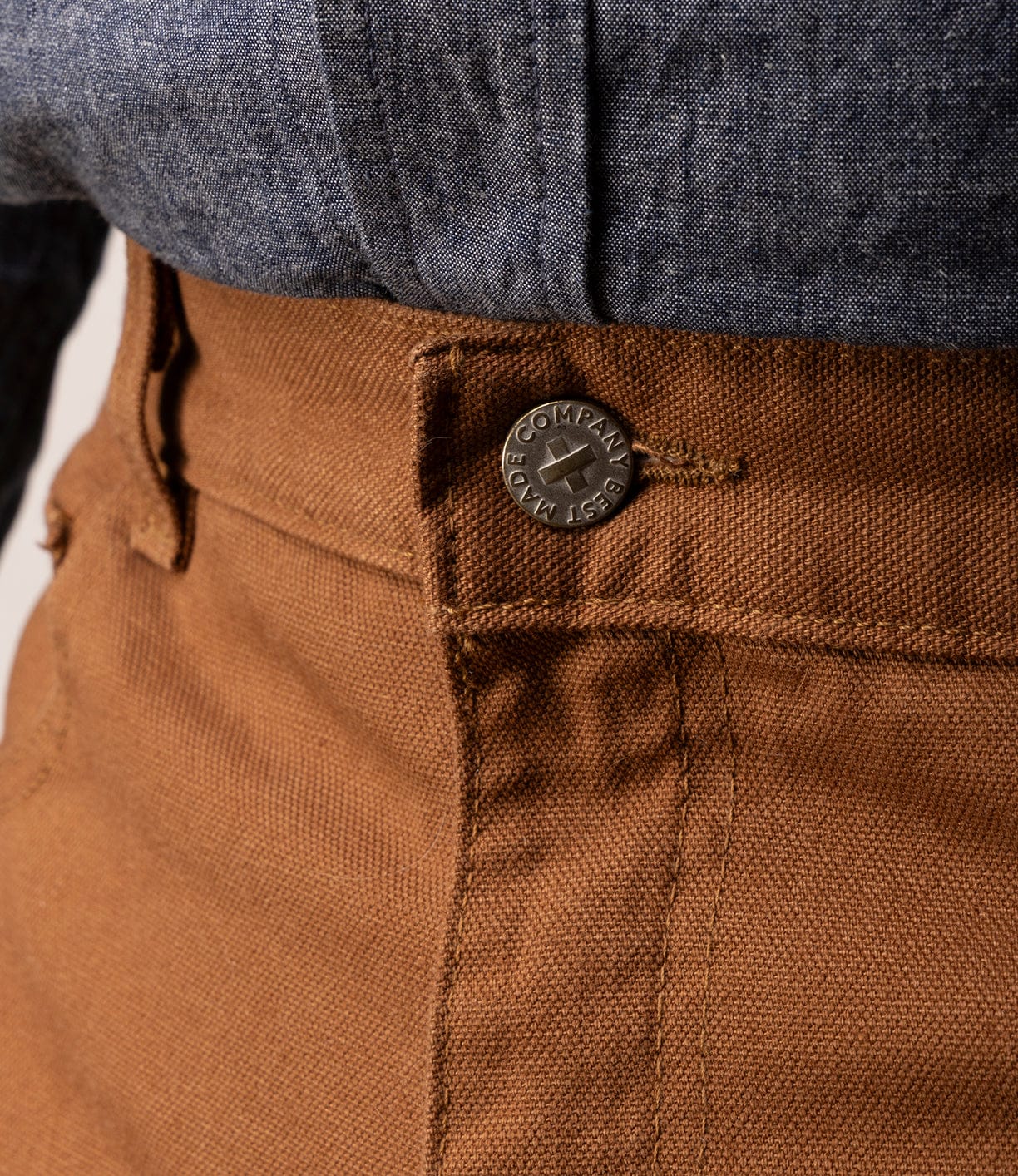 Close-up of a person wearing Best Made Co's Standard 5-Pocket Pant M2 Field Brown, featuring heavyweight cotton with a textured finish and metal button, paired with a dark blue chambray shirt tucked in. Focus is on the waistband and button.
