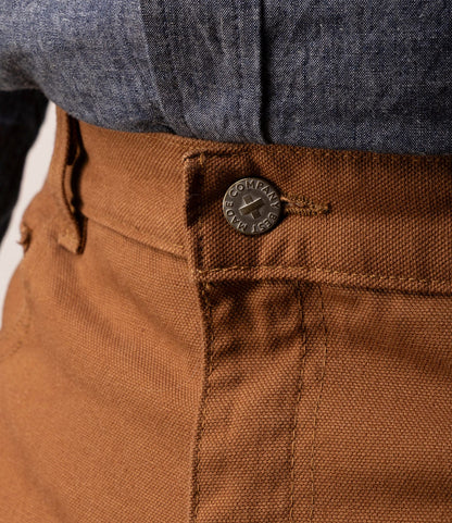 Close-up of a person wearing Best Made Co. Standard 5-Pocket Pant M2 Field Brown with an engraved metal button and blue-gray shirt tucked in. The classic straight cut and heavyweight cotton highlight the detailed seams.