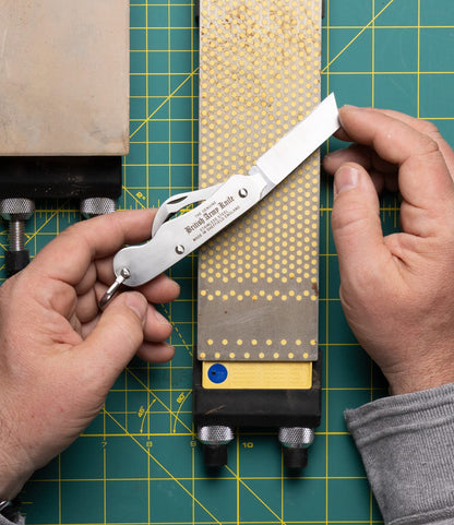 A person sharpens the Best Made Co. British Army Knife on a rectangular stone, holding the utility knife at an angle with both hands over a gridded cutting mat.