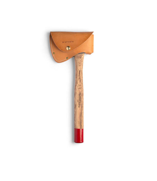 A person in a brown jacket holds Best Made Co.'s Spark (Straight-Hold Edition), featuring an Appalachian hickory handle and a red-tipped high-carbon steel axe head, sheathed in tan leather reading BE OPTIMISTIC. Natural window light fills the scene.