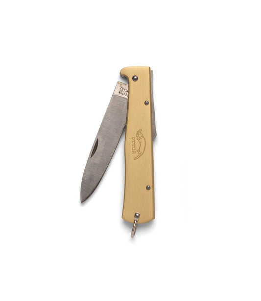 A person holds an open Best Made Co Brass Mercator pocket knife with a cream handle against a sharpening stone, placed on a green cutting mat with measurement lines and nearby tools visible.