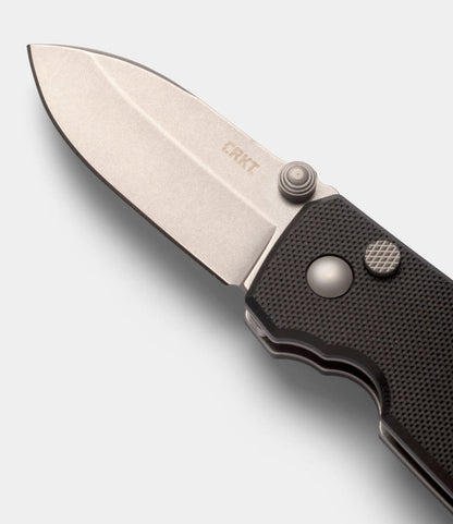 A close-up of the Best Made Co. CRKT Squid pocket knife shows its D2 steel blade, textured black handle, CRKT logo on the blade, thumb stud for easy opening, and secure button lock mechanism.