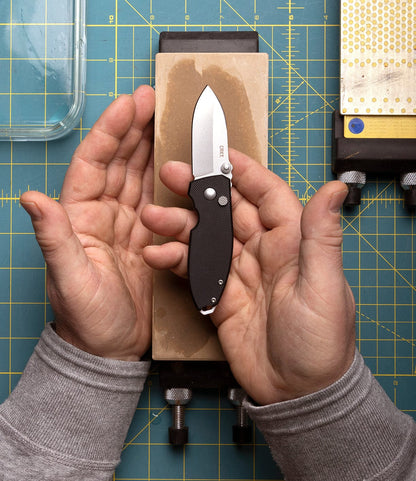 A person holds a Best Made Co. CRKT Squid pocket knife above a sharpening stone, both hands framing the D2 steel blade. In the background is a green grid-patterned cutting mat with various sharpening tools.