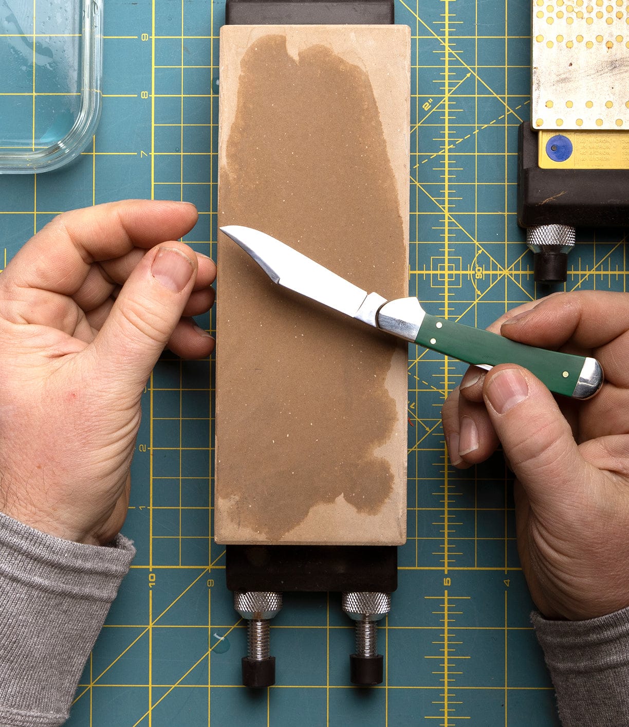 A person holds a Best Made Co Mini Copperlock knife above a whetstone, ready to sharpen the blade on a green cutting mat with yellow grid lines.