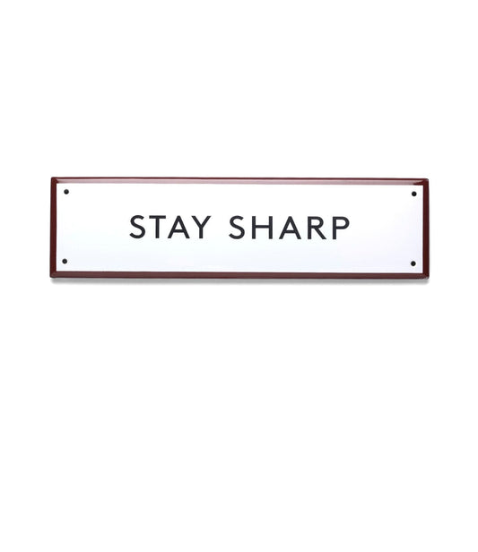 Axes and hammers with wooden handles stand upright by a rustic wall. Below, a white “Stay Sharp” Enamel Steel Sign by Best Made Company Co. displays the bold message in black letters, hand-crafted in Poland.