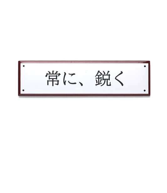 A wooden wall features five Japanese blades, a folding pocket knife, sharpener, and yellow tag below a magnetic strip. Above them hangs Best Made Company Co.’s “Stay Sharp” Enamel Steel Sign (Kanji Edition).