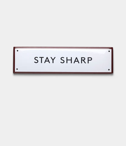 The Best Made Co. “Stay Sharp” Enamel Steel Sign features black text on a rectangular white enamel surface with a thin dark border, showcased against a plain light gray background for a classic, durable look.