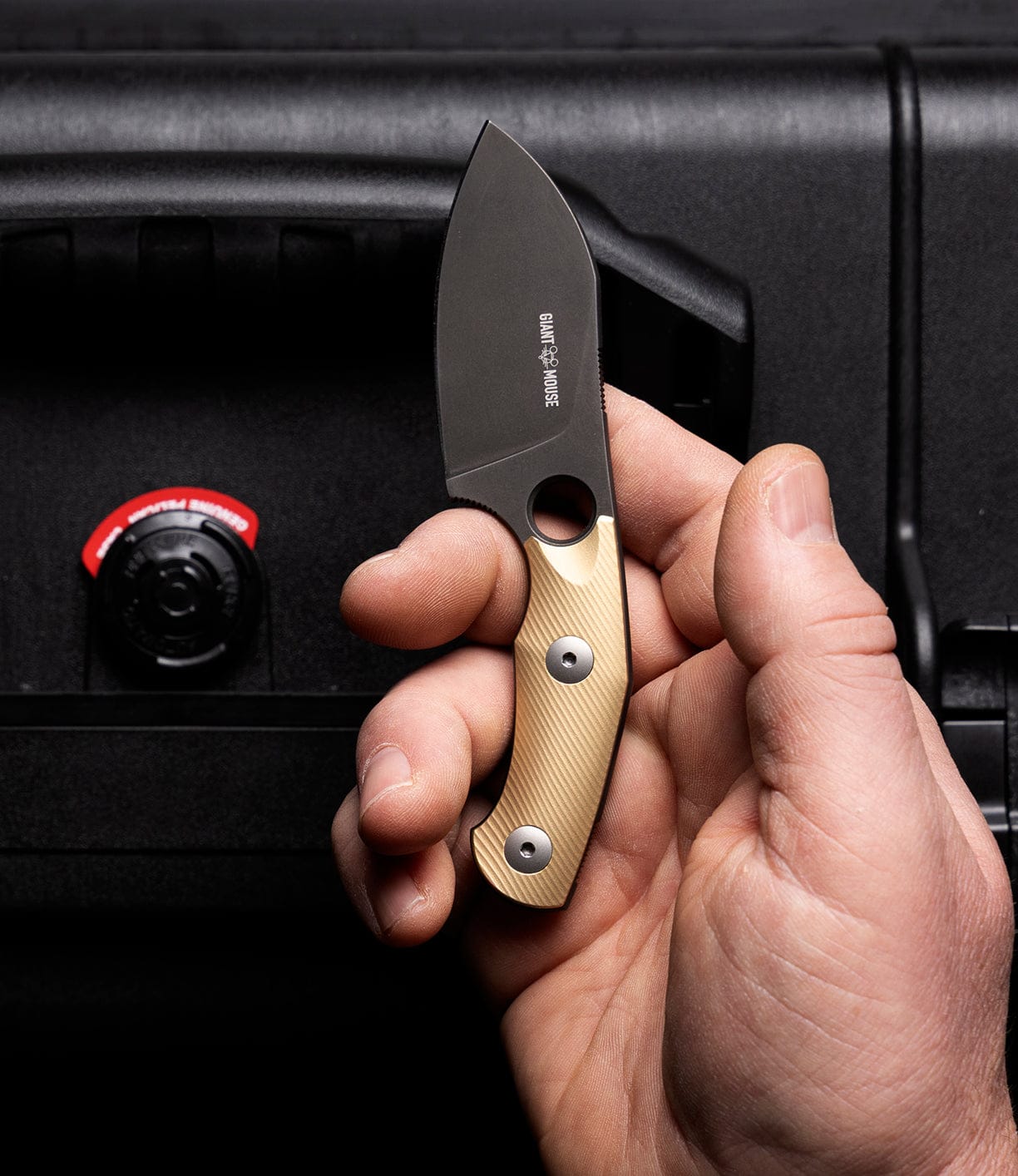 A hand holds the Best Made Co GiantMouse GMF1-P (BMC Edition) fixed-blade knife with a gold and black handle, in front of a black case with a red and black locking mechanism.