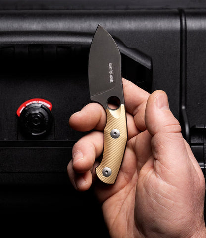 A hand holds the Best Made Co. GiantMouse GMF1-P (BMC Edition) folding knife with brass scales and a black blade, shown with its handmade leather sheath and a black case featuring a red and black knob in the background.