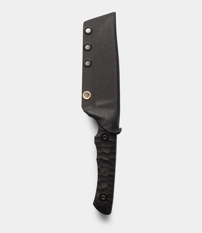 The NWBW Field Knife (BMC Edition) by Best Made Co. features black D2 tool steel, a textured handle, and a fitted black sheath with three metal rivets and one brass ring, shown on a light gray background.
