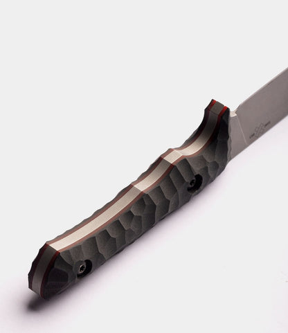A close-up of a Best Made Co. NWBW Field Knife (BMC Edition) handle with a textured, hammered metal design and visible screws, set against a white background. The D2 tool steel blade is partially visible on the right.