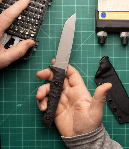 A person holds a Best Made Co. NWBW Field Knife (BMC Edition) with a textured black handle over a green cutting mat. Nearby are a screwdriver set, sheath, and sharpener, highlighting quality D2 tool steel craftsmanship.
