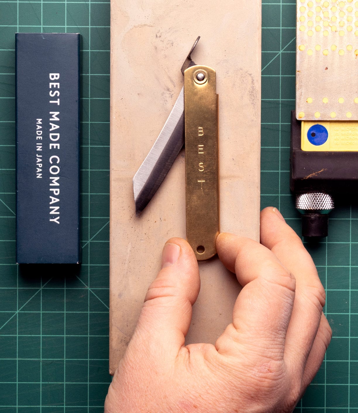 A hand holds the Max Brass Higo Knife by Best Made Co, engraved with MUST, resting on a sharpening stone. Nearby are a Best Made Co box and a sharpening tool on a green cutting mat.