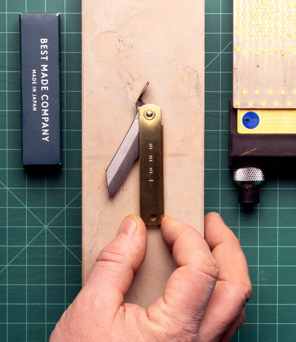 A hand holds a partially open Mid Brass Higo Knife from Best Made Co on a sharpening stone beside its branded box and a metal tool, all resting on a green cutting mat.