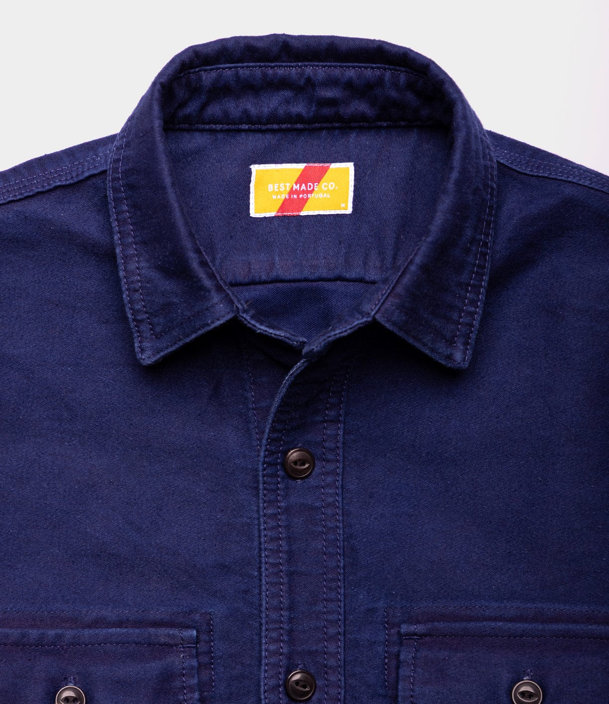 Close-up of the Best Made Co Indigo Moleskin Work Shirt, featuring a collar, two front pockets, and a yellow and red label sewn inside the neck, neatly arranged against a light background.