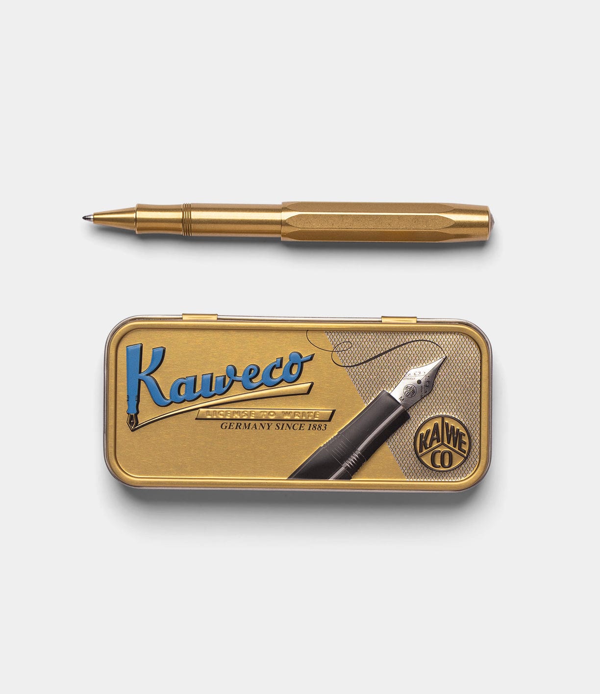 A Best Made Co. Kaweco Sport brass pocket pen with its cap off rests atop a vintage-style gold tin box with blue and black logo lettering and a nib illustration. Uses the G2 rollerball refill for smooth writing.