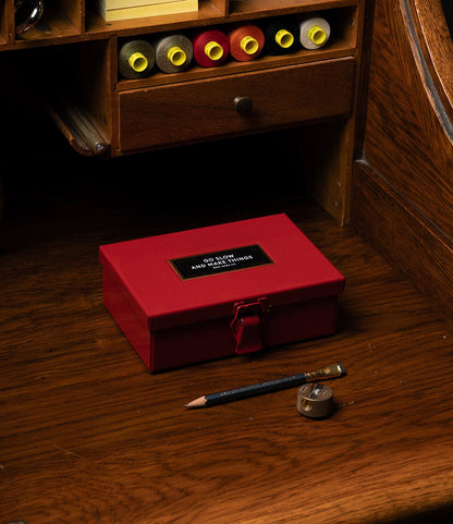 A red Best Made Co. Type 6 Strongbox, Go Slow Edition, sits on a wooden desk with a pencil and sharpener nearby. Above are wooden shelves holding rolls of colored paper.