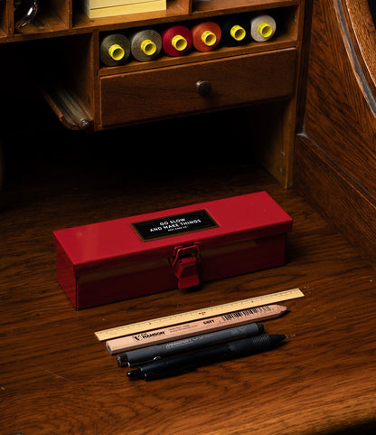 A red steel Type P Strongbox, Go Slow Edition by Best Made Co., sits on a wooden desk with a ruler, pencil, pen, and marker in front. On shelves above are papers and yellow, red, and gray cylinders. Limited edition.