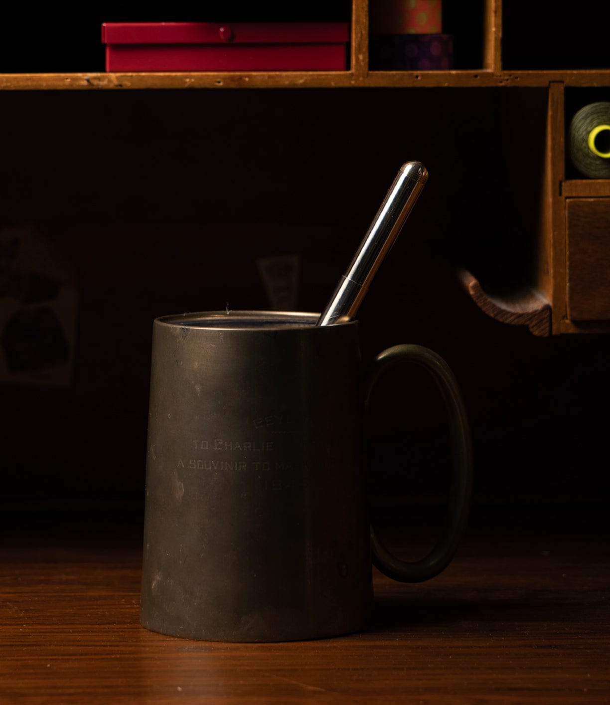 A dark metal mug with a handle rests on wood, holding a Kaweco Supra Fountain Pen by Best Made Co. The dimly lit background shows a wooden shelf lined with small objects and boxes.