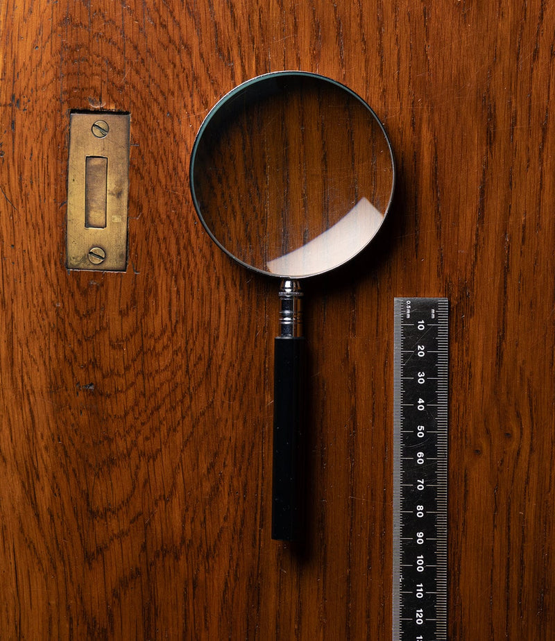 A magnifying glass and a black ruler are placed on a wooden surface near a brass door plate. The wood has a visible grain pattern.