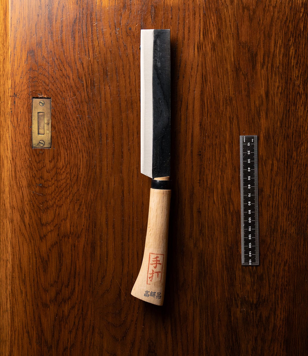 A Japanese Hatchet by Best Made Co, featuring a wooden handle with red Japanese characters and a black blade, rests on a wooden surface beside a black ruler.