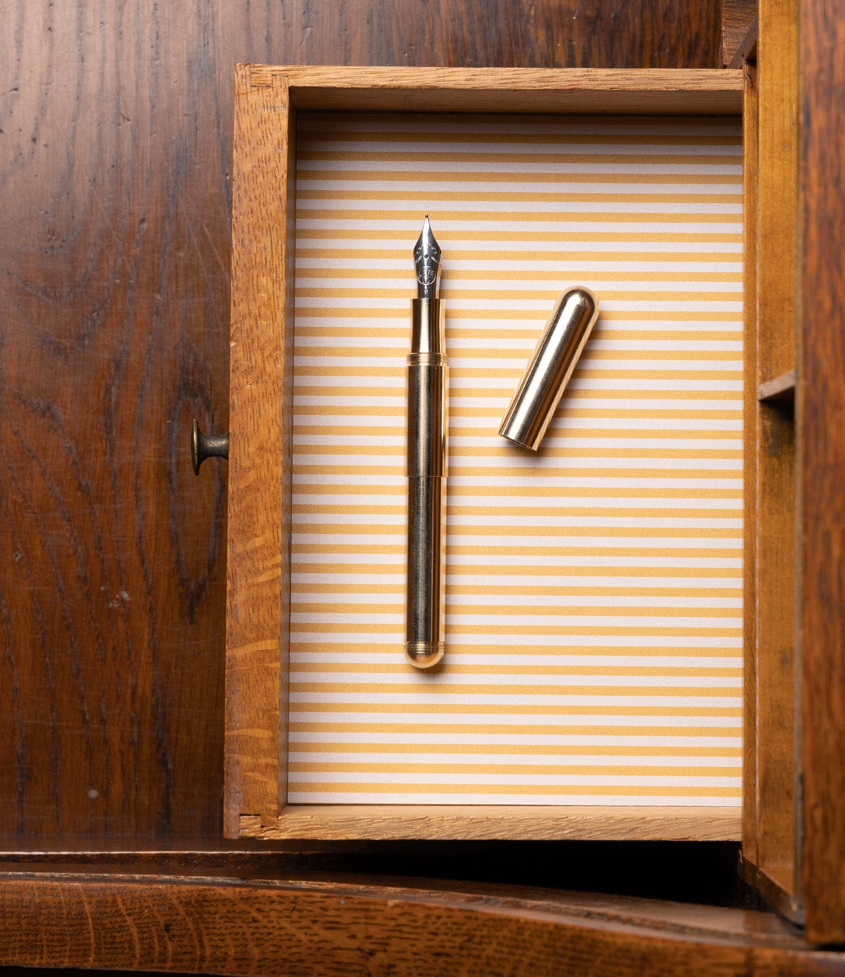 A Best Made Co. Kaweco Supra Fountain Pen and its cap rest inside a wooden drawer lined with yellow and white striped paper, partially open within a wooden cabinet.