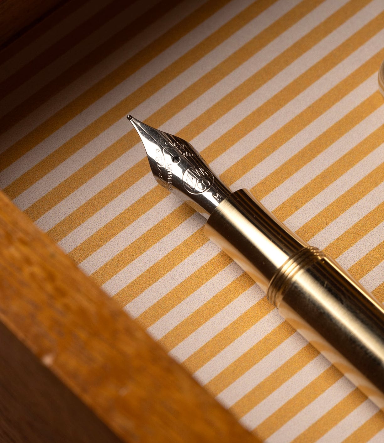 A close-up of a Kaweco Supra Fountain Pen nib by Best Made Co., resting on a striped yellow and white surface, with part of a wooden box visible.