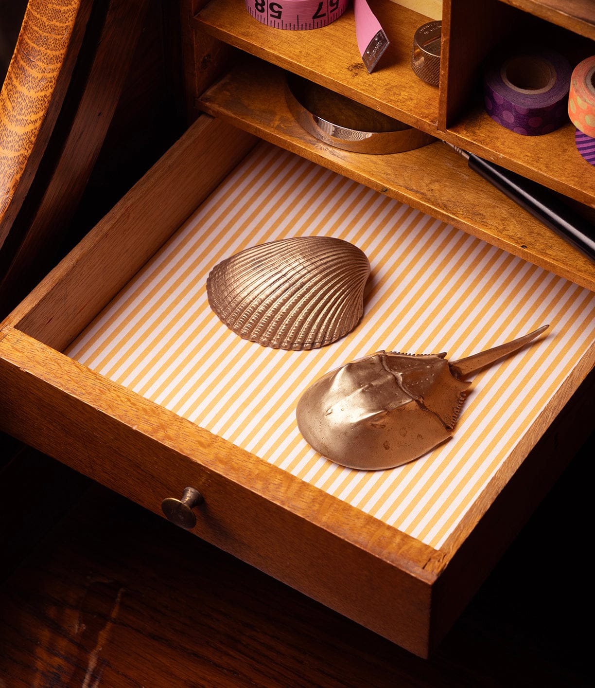 A wooden drawer lined with yellow and white striped paper holds a metallic seashell and the Best Made Co. Horseshoe Crab Opener. The open drawer is part of a desk topped with various stationery items.