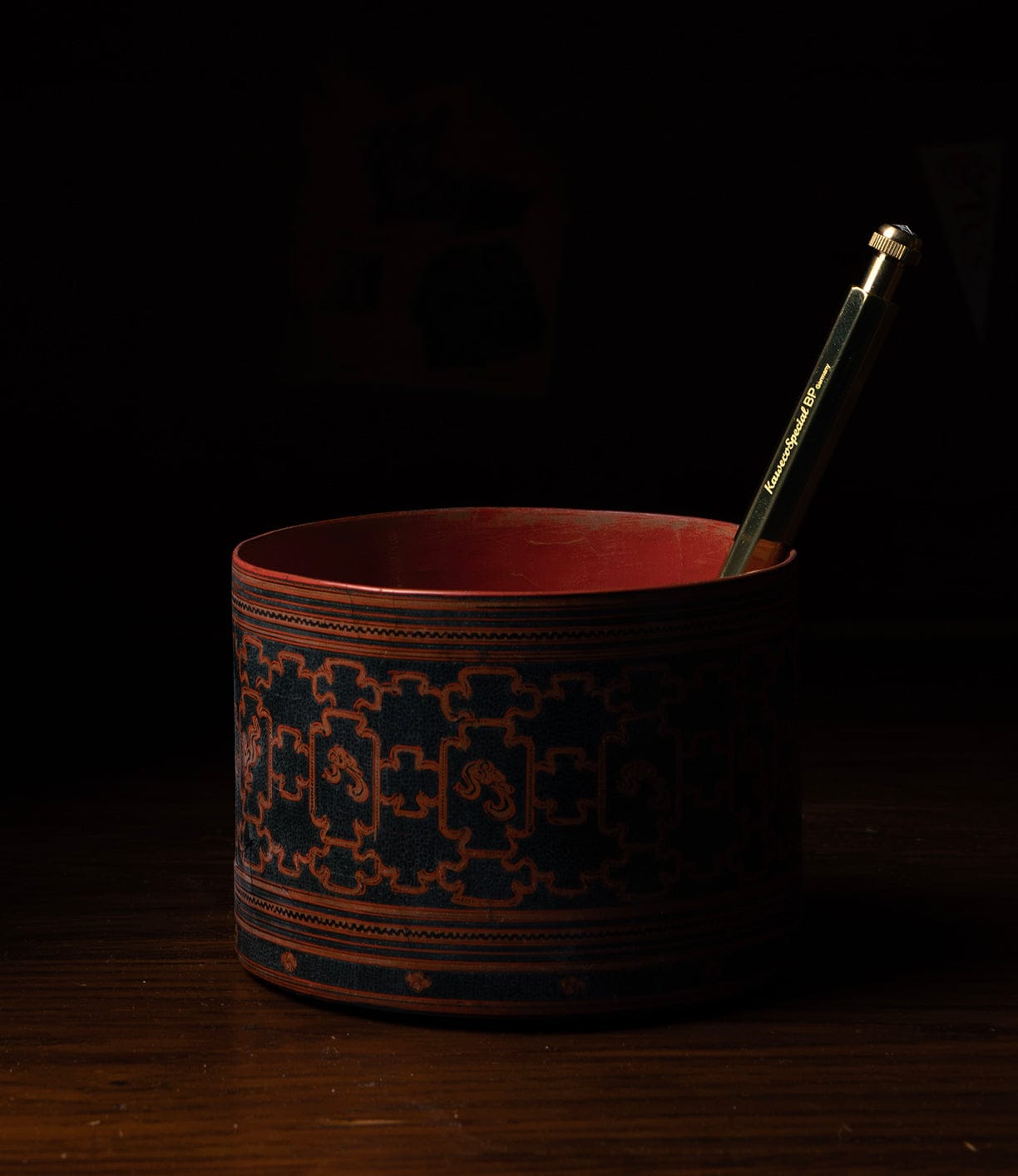 A Kaweco Special green pencil and Best Made Co. brass ballpoint pen rest in an ornate, octagonal holder on a wooden surface, set against a dark background.