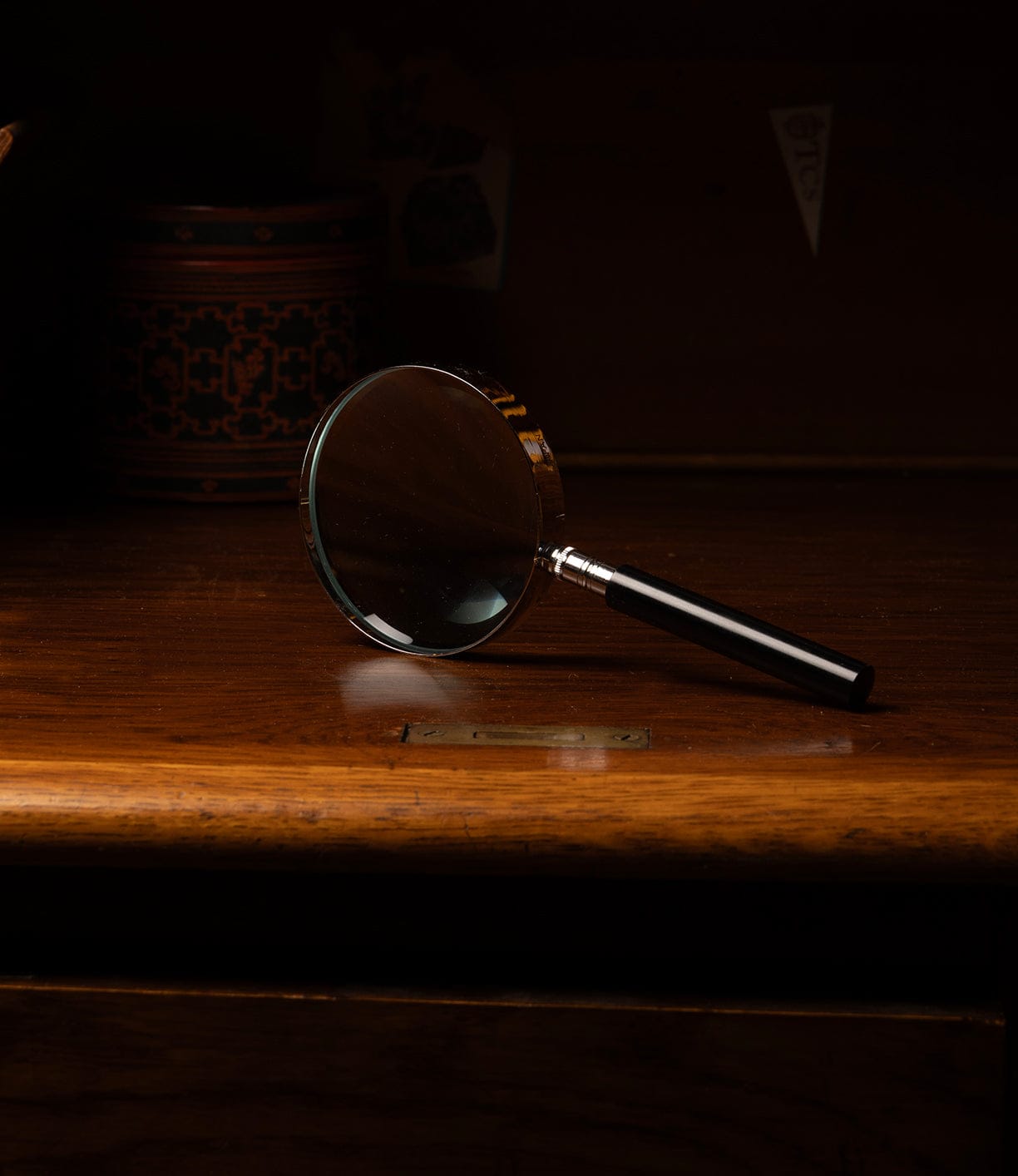 The Best Made Co. Japanese Magnifying Glass, featuring precision optics and a black handle, sits on a polished wooden surface with a decorative container nearby and a dark backdrop enhancing the scene.