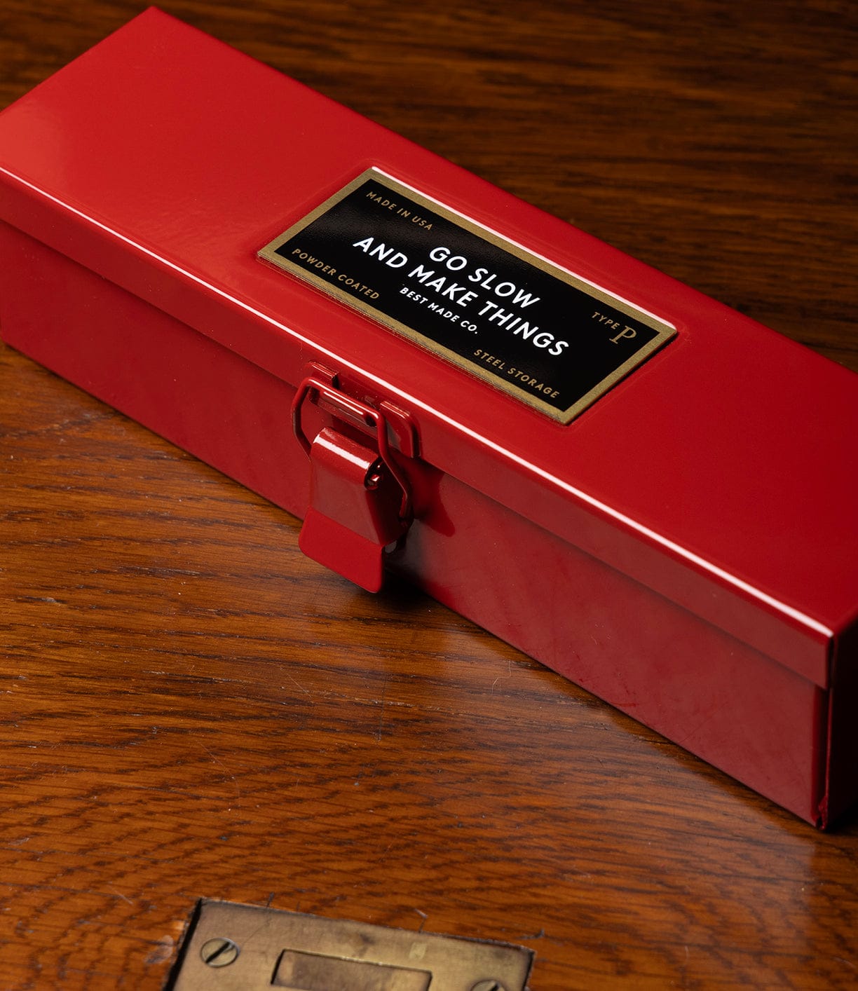 A Best Made Co Type P Strongbox, Go Slow Edition—a red steel pencil storage box with a latch—sits on a wooden surface. Its label reads, GO SLOW AND MAKE THINGS.