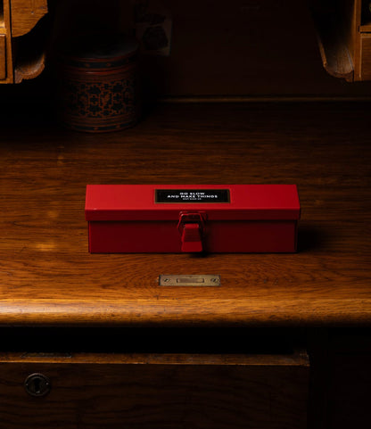 A red rectangular Best Made Co. Type P Strongbox, Go Slow Edition with a clasp rests on a polished wooden desk. The box features a black and white label on top, with background items partially visible.