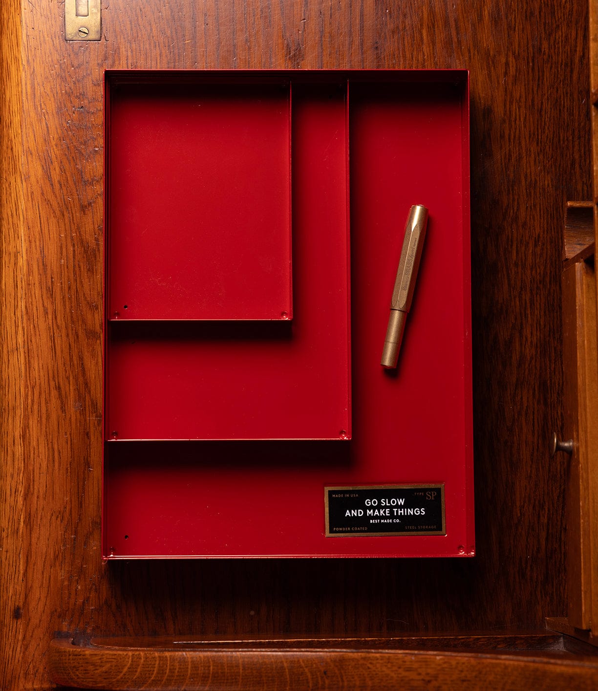 A red Spare Parts Tray from Best Made Co’s "Go Slow Edition" sits on wood, holding a gold pen and a plaque reading “GO SLOW AND MAKE THINGS.”.