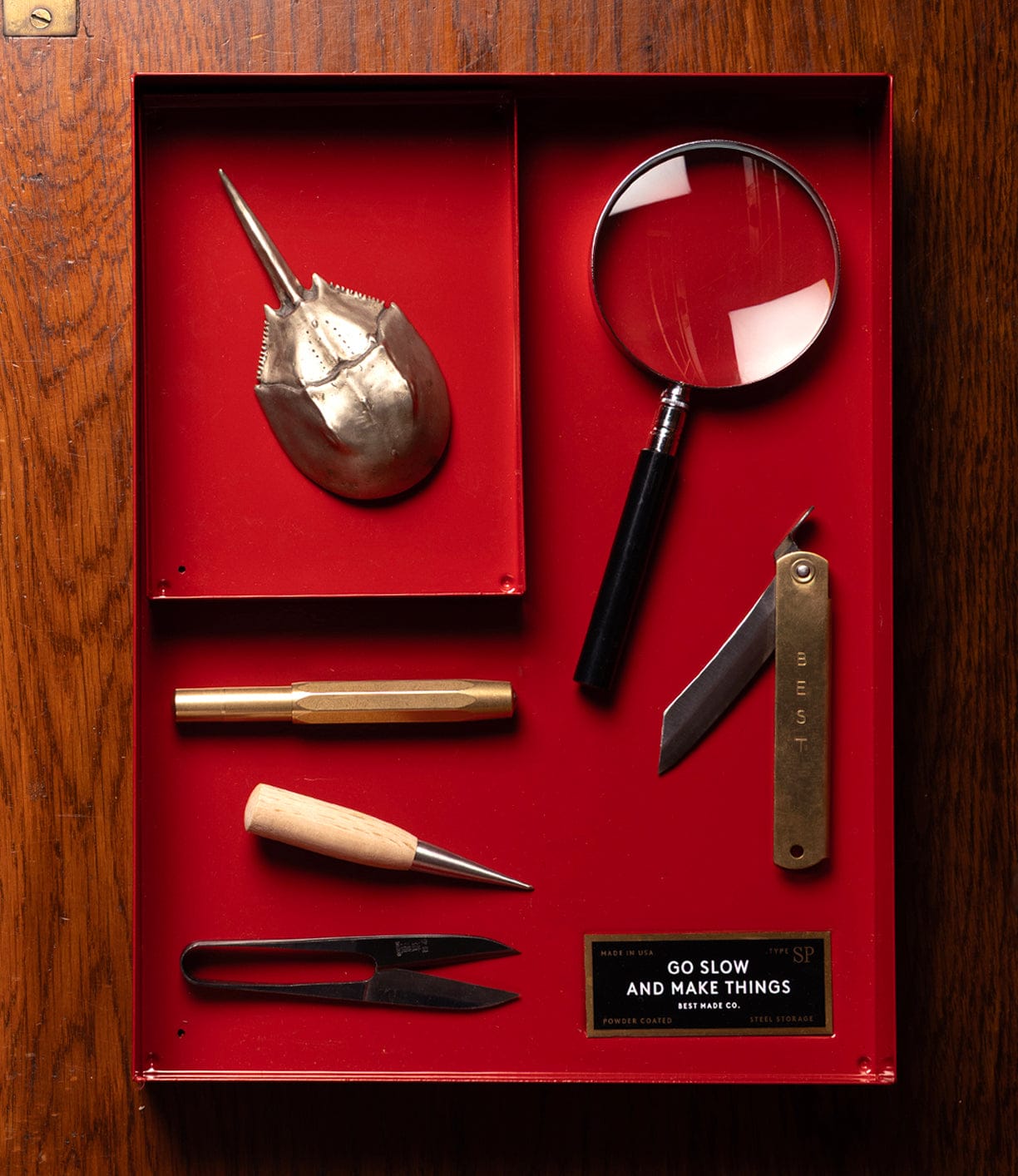 A red tray on a wooden surface holds a pocket knife, a boxed item, a yellow circular object, a seashell, a horseshoe crab shell, a magnifying glass, two pens, tweezers, a pen knife, and a label.