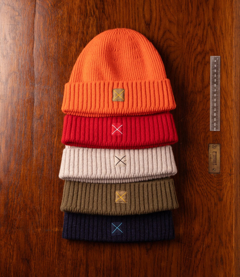 Five ribbed knit beanies in orange, red, beige, olive, and navy are stacked on a wooden surface next to a clear ruler and a brass keyhole plate. Each beanie has a small patch with an embroidered X.