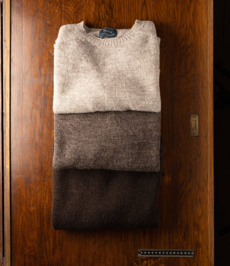 A neatly folded stack of three wool sweaters in beige, brown, and dark brown lies on a wooden surface with visible grain and a metal lock.