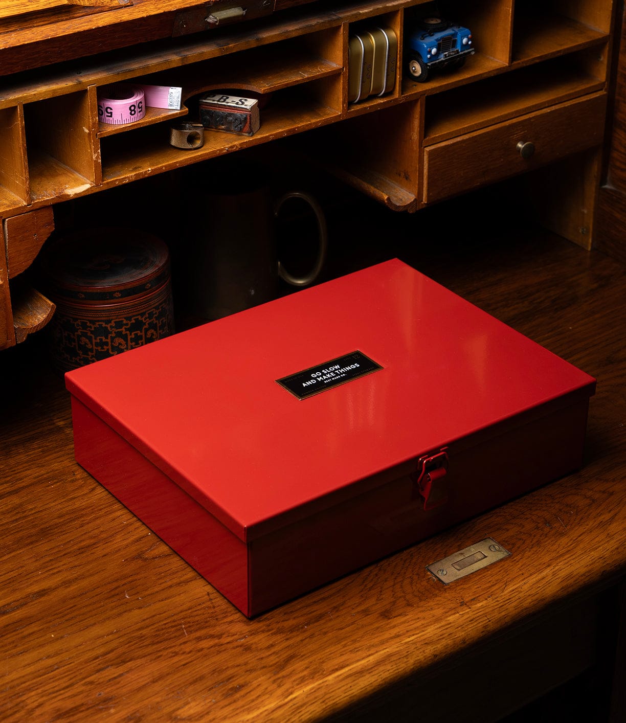 A closed, shiny red Best Made Co Type 12 Strongbox, Go Slow Edition with a black label rests on a warm vintage wooden desk beneath cubbyholes filled with small items.