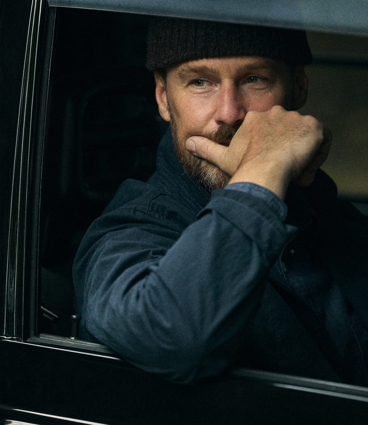 A man with a beard and dark beanie sits in a car, resting his arm on the window frame and his hand on his chin, looking thoughtful in the Possibilities Jacket Navy by Best Made Co.