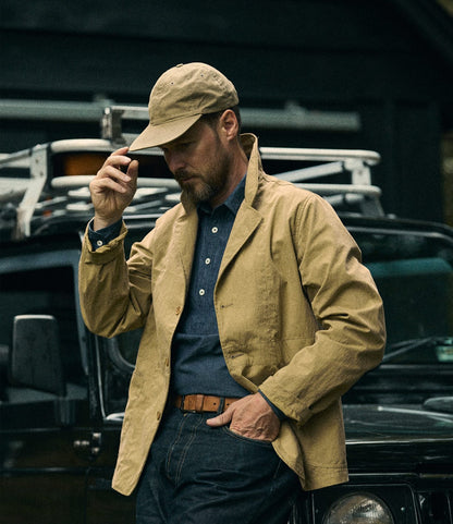 A man dressed casually in a Best Made Co. Possibilities Jacket in tan and a cap stands in front of a black vehicle, adjusting his hat with one hand and looking down, wearing a blue shirt and brown belt.