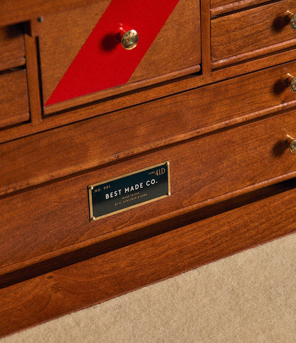 Close-up of the Gerstner 41D Founder’s Edition wooden cabinet by Best Made Co., featuring brass knobs, a distinctive red diagonal stripe, and a metal Best Made Co. label. The cabinet rests on a beige carpet.