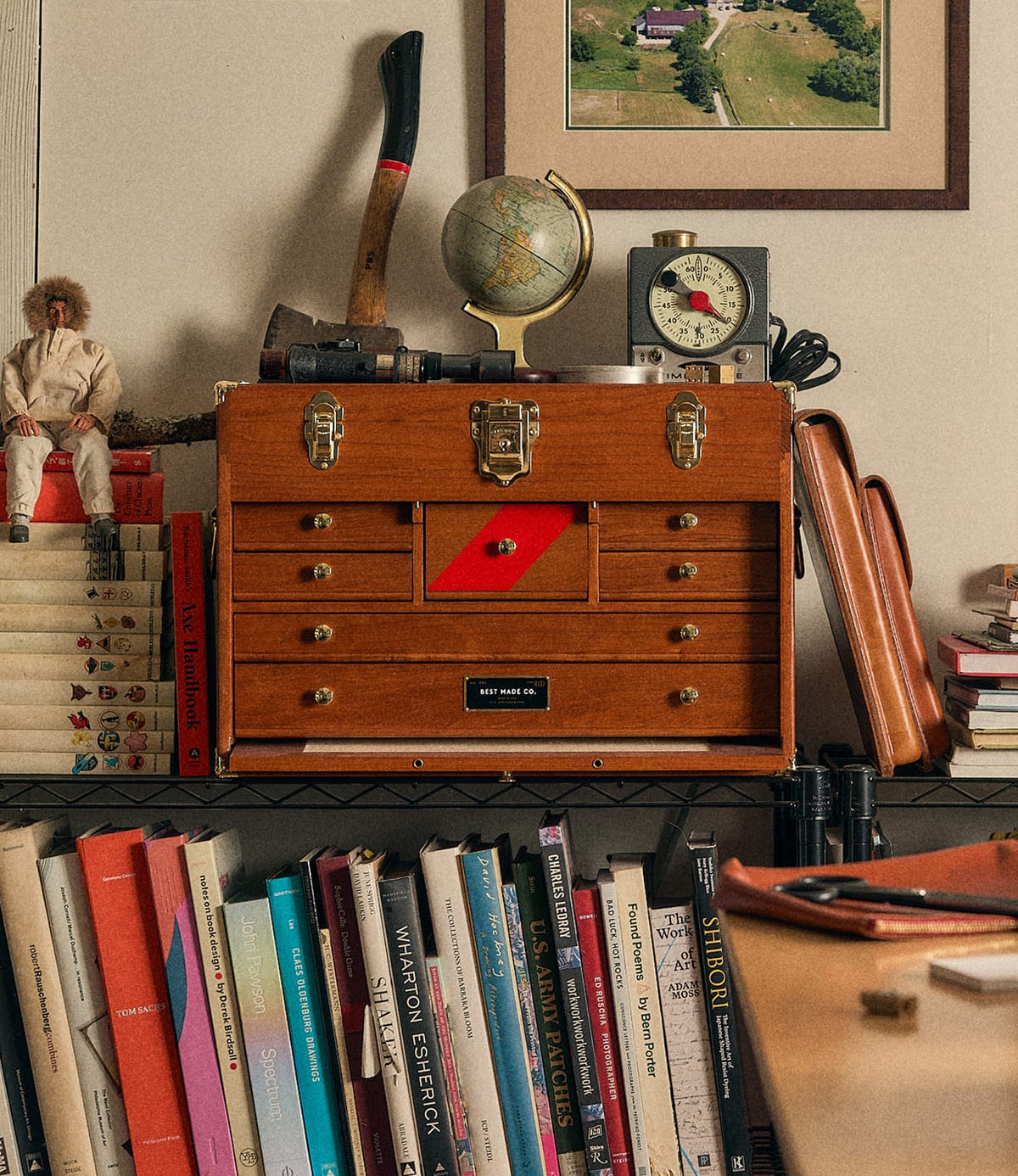 A Best Made Co. Gerstner 41D Founder’s Edition machinist chest sits on a shelf surrounded by books, a globe, an axe, a clock, and a doll, with a framed aerial photo hanging on the wall above.