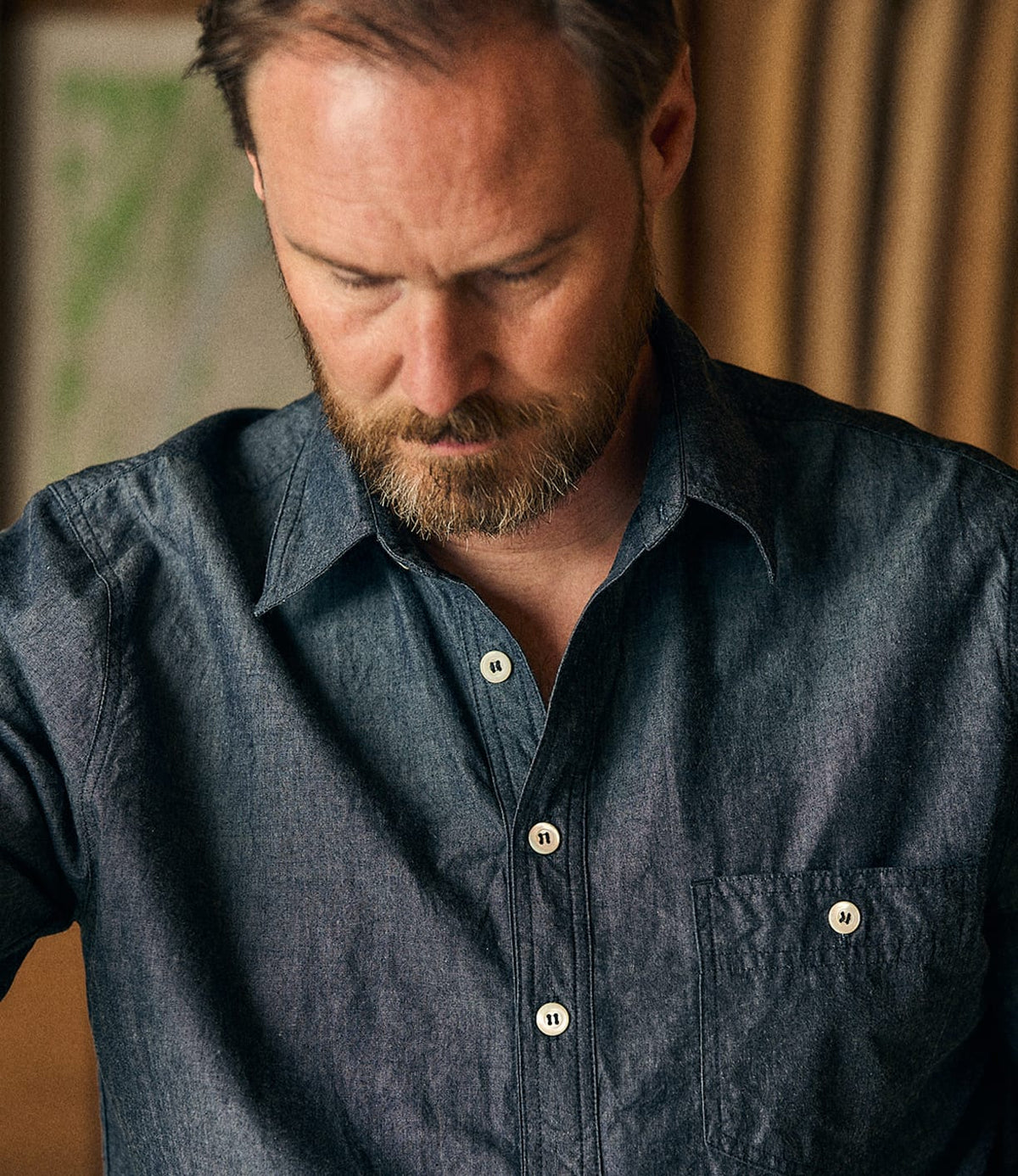 A man with a neatly trimmed beard looks down, wearing the Best Made Co. Indigo Chambray Popover, expertly crafted from rope-dyed denim. The softly blurred background features warm tones and indistinct shapes.