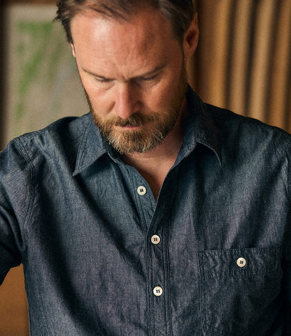 A man with a neatly trimmed beard looks down, wearing the Best Made Co. Indigo Chambray Popover, expertly crafted from rope-dyed denim. The softly blurred background features warm tones and indistinct shapes.