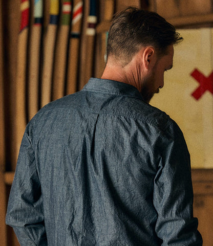 A man with short hair, seen from behind, wears the Best Made Co. Indigo Chambray Popover. Colorful oars and a flag featuring a red X appear blurred in the background.